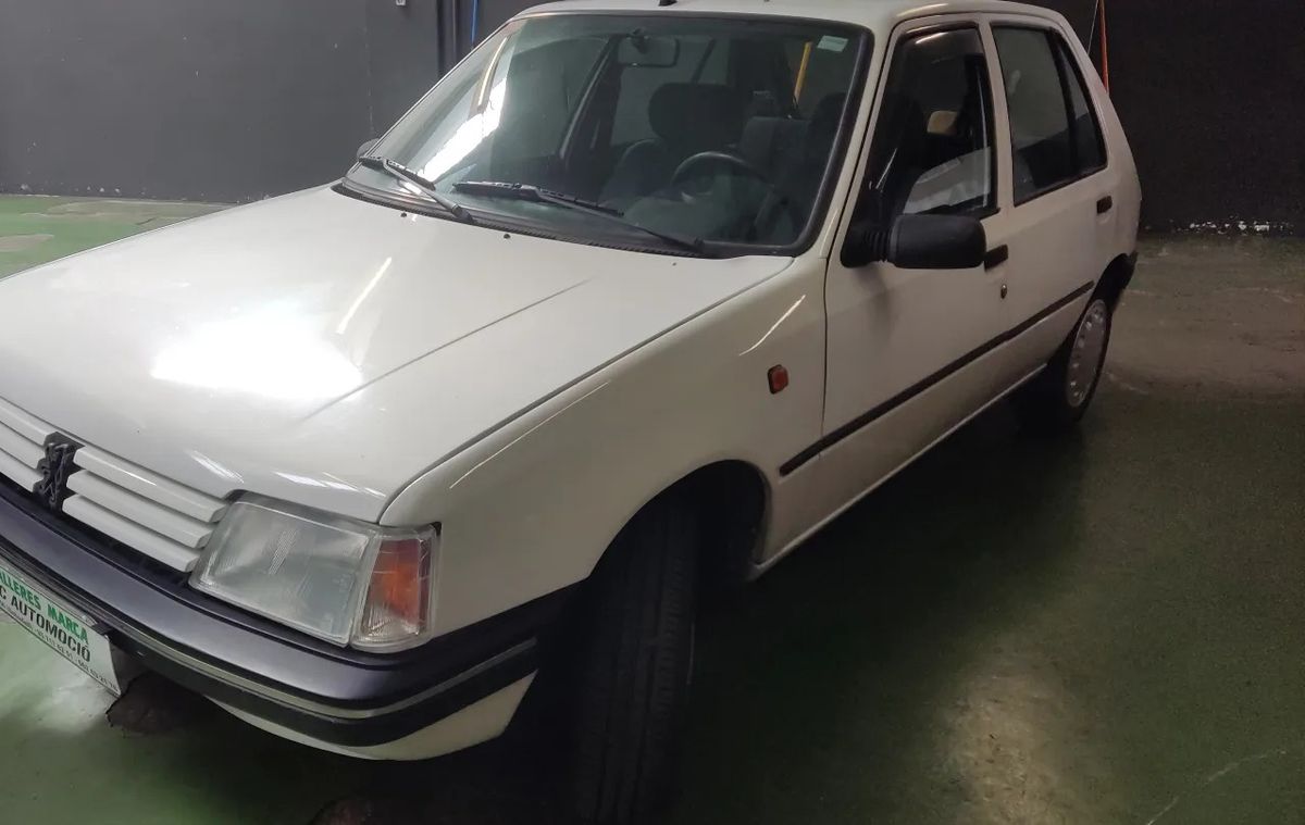 Peugeot 205 blanco sobre el suelo verde del taller, con la funda de matrícula de Talleres Marca